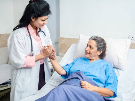 Smiling Indian caring doctor supporting holding hand of olde senior female patient lying on bed at clinic or hospital. Elderly people health care concept.
